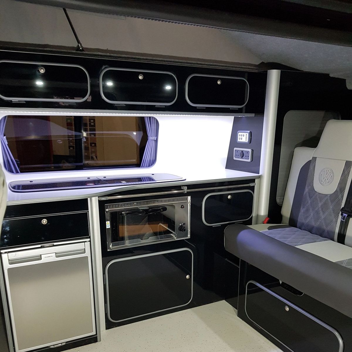Black gloss kitchen units with a fridge, grill and LED strip lighting beside a bench seat